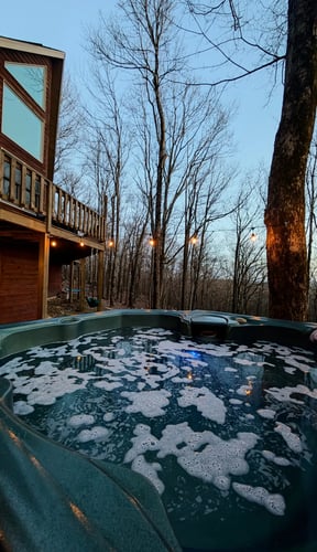 Bañera de hidromasaje al aire libre junto a una cabaña al anochecer con guirnaldas de luces y árboles sin hojas en invierno